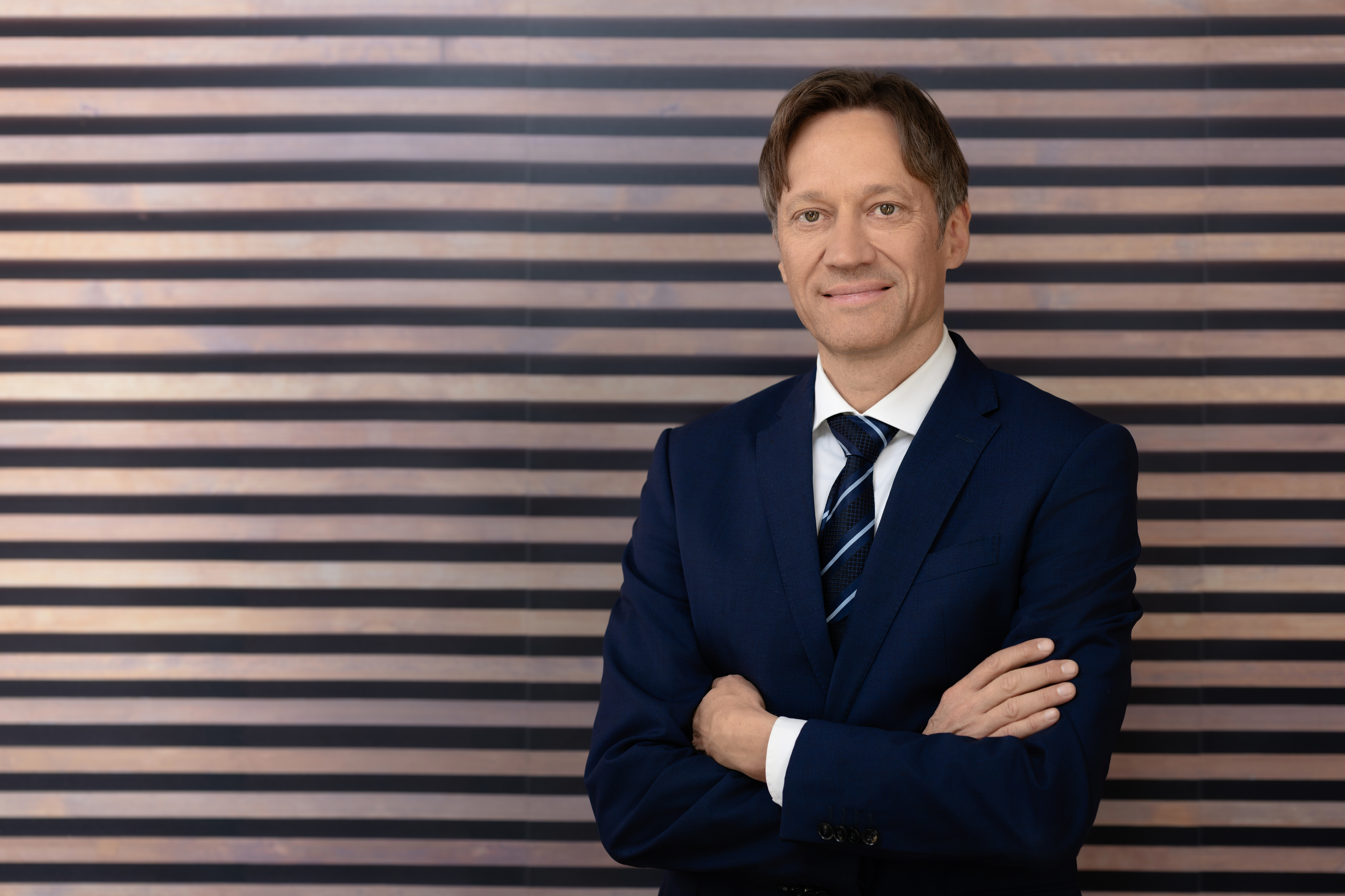 Der CEO von der Firma Siemens Mobility, Herr Michael Peter, steht mit verschränkten Armen und lächelnd vor einer Holzlamellenwand Der CEO von der Firma Siemens Mobility, Herr Michael Peter, steht mit verschränkten Armen und lächelnd vor einer Holzlamellenwand